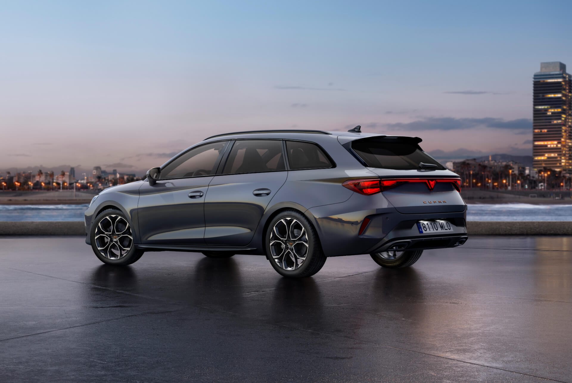 Heckansicht des neuen CUPRA Leon Sportstourer 2024 in Glossy Grey mit dynamischer Fahrwerksteuerung (DCC) sowie schwarz-kupferfarbenen Leichtmetallrädern. Im Hintergrund die Stadt beim Sonnenuntergang.