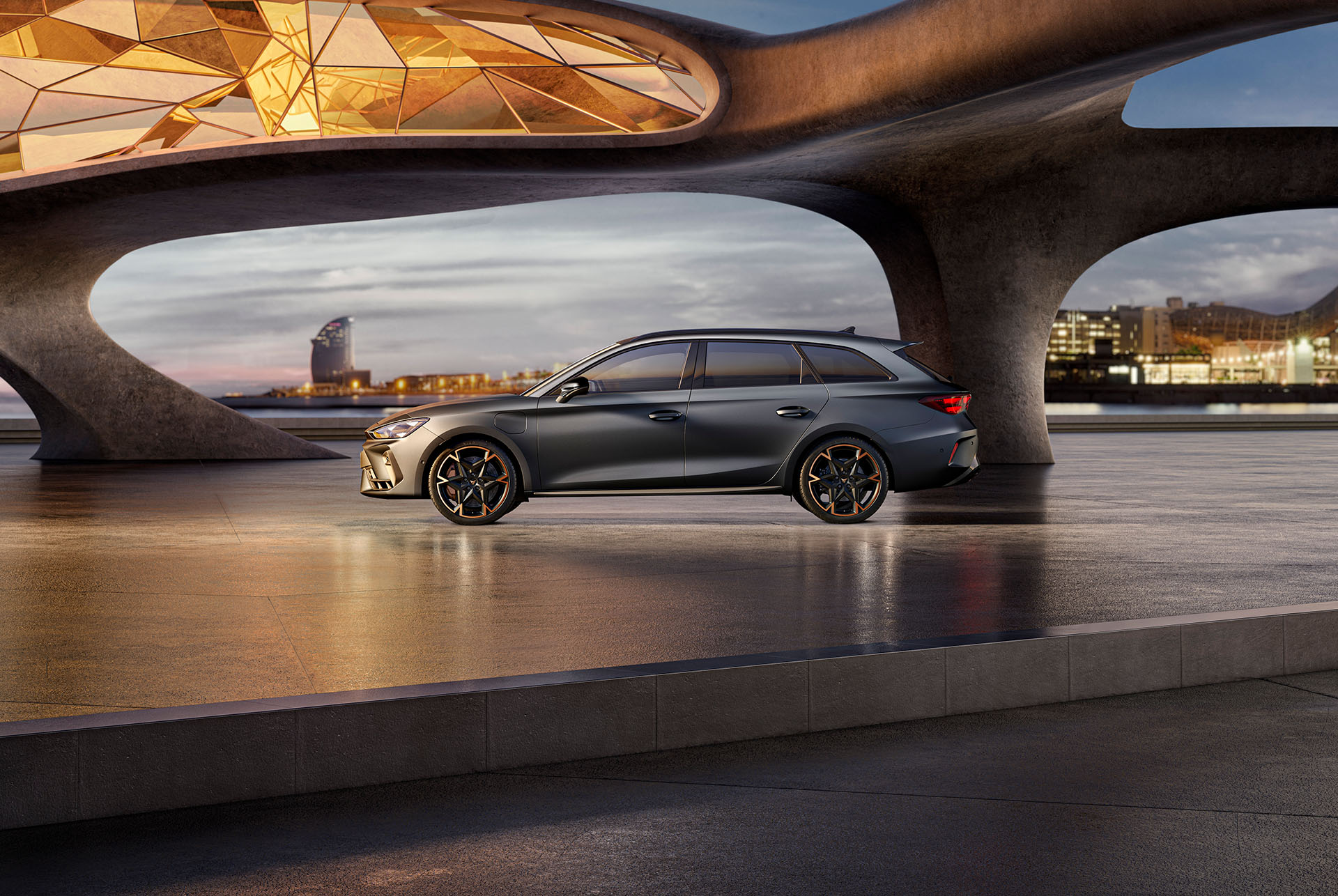 Neue CUPRA Leon Sportstourer 2024 Fahrzeuge und Ausstattung, parkiert auf Beton, Struktur mit leuchtenden Kanten, Blick auf die Skyline der Stadt bei Sonnenuntergang. Das Fahrzeug wird in der Farbe Matte Grey mit kupferfarbenen und schwarzen Leichtmetallrädern präsentiert.