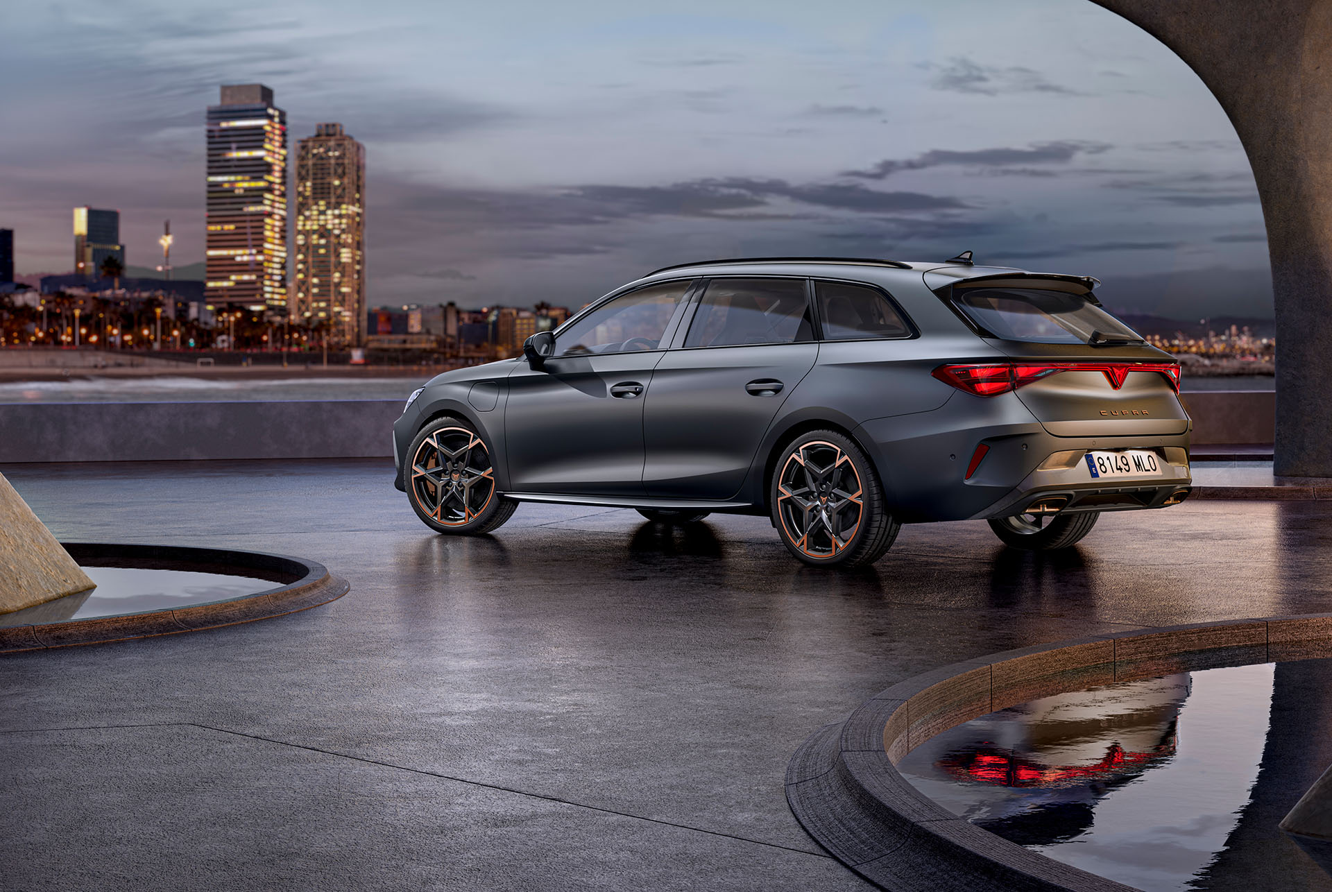 Technologie de la nouvelle CUPRA Leon Sportstourer, garée sur du béton, structure aux contours lumineux, vue sur l’horizon de la ville au crépuscule. Le véhicule est présenté en couleur Matte Grey avec des jantes en alliage léger cuivrées et noires.
