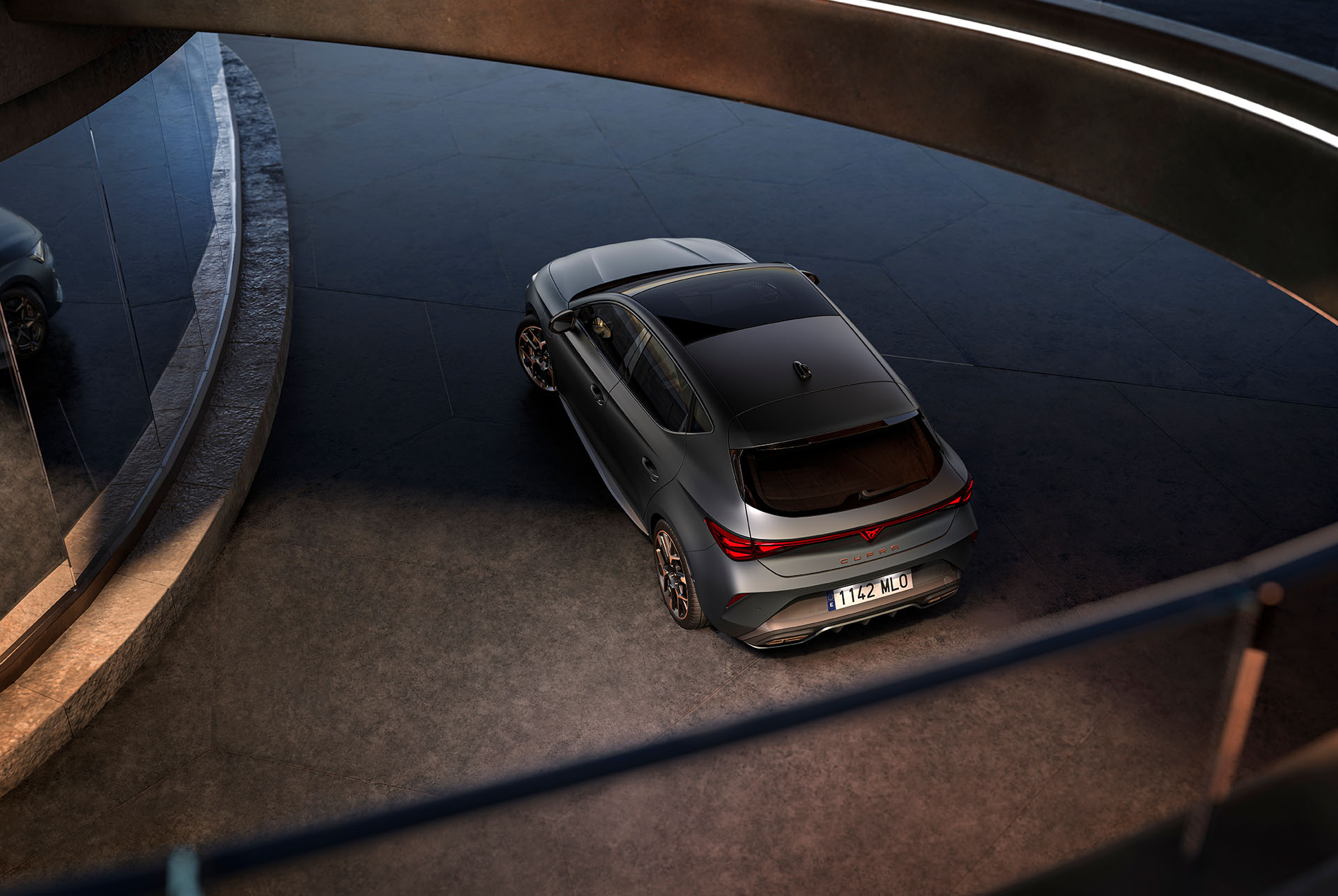 Hybridauto-Technologie im neuen CUPRA Leon 2024, LED-Heckleuchten, PHEV-Fahrzeug auf Beton parkiert, Struktur mit leuchtenden Kanten. Das Fahrzeug ist in mattgrauem Finish mit Schmiedefelgen in Kupfer und Schwarz abgebildet.