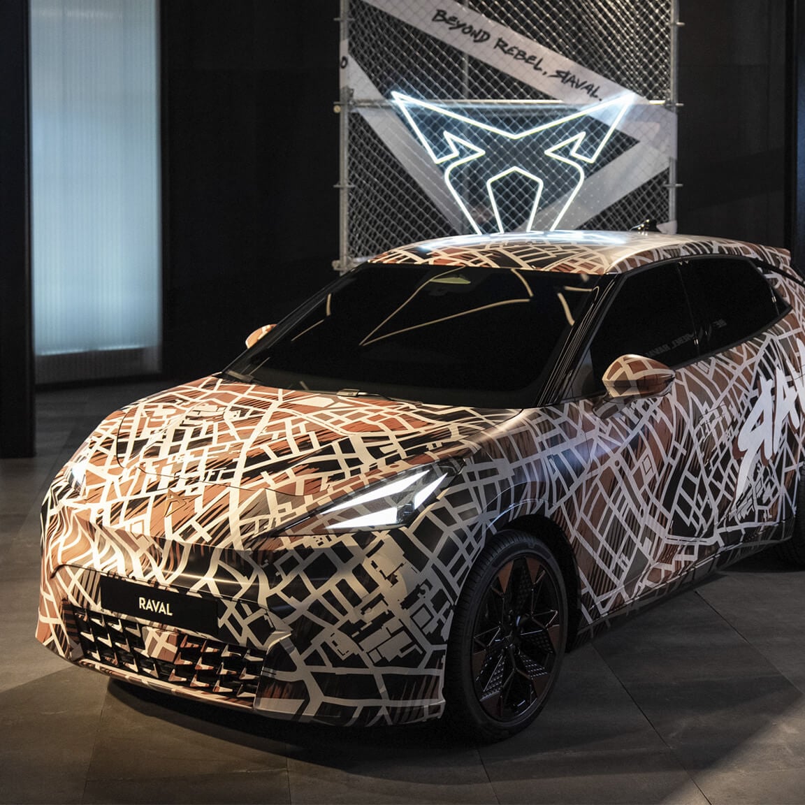  La CUPRA Raval, recouverte d’une enveloppe de camouflage marron et blanc, est exposée à l’intérieur. Derrière la voiture se trouve un panneau de clôture à maillons de chaîne avec un logo CUPRA blanc étincelant et la mention « BEYOND REBEL, RAVAL. ».
