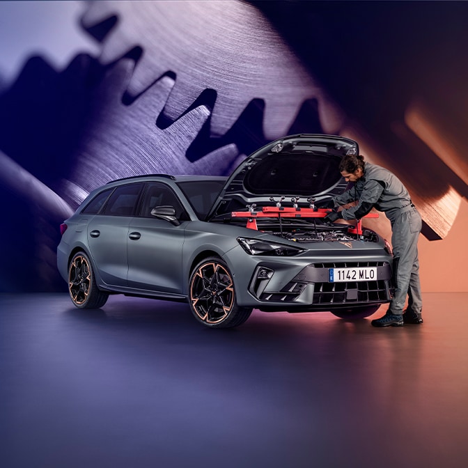 Ein Techniker in einem grauen Overall untersucht den Motorraum eines CUPRA Terramar in einem Studio; im Hintergrund ist ein grosser Bildschirm zu sehen, der eine Nahaufnahme von metallischen Zahnrädern zeigt.