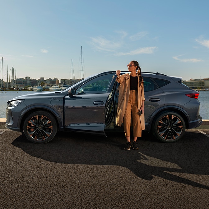 Ein sonnendurchflutetes Foto einer Frau in einem Trenchcoat, die ein Smartphone hält und nach rechts schaut. Sie steht neben der offenen Fahrertür eines dunkelgrauen CUPRA-Autos, das an einem Yachthafen mit Blick auf die Stadt geparkt ist.