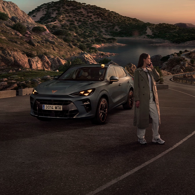 Ein Weitwinkel-Landschaftsbild bei Dämmerung, das eine Frau in einem Trenchcoat zeigt, die neben einem CUPRA Terramar auf einer kurvigen Küstenstrasse steht, mit felsigen Hügeln und einem ruhigen Meer im Hintergrund.