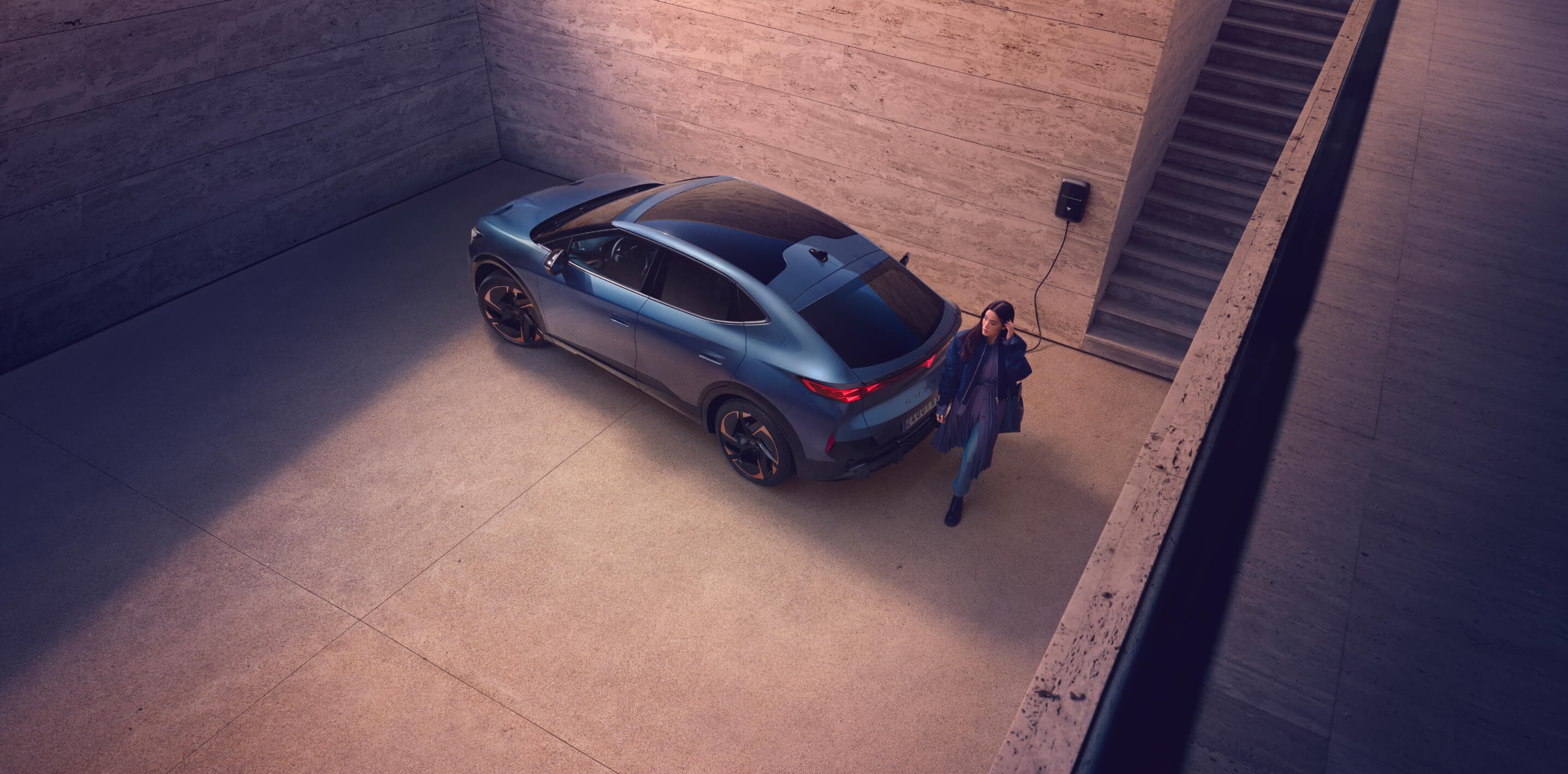 elektrischer SUV, der in eine Wallbox in einer minimalistischen Betonauffahrt gesteckt wird, Fahrer aus dem Auto, präsentiert schlankes Automobildesign und eine Ladestation für Elektrofahrzeuge